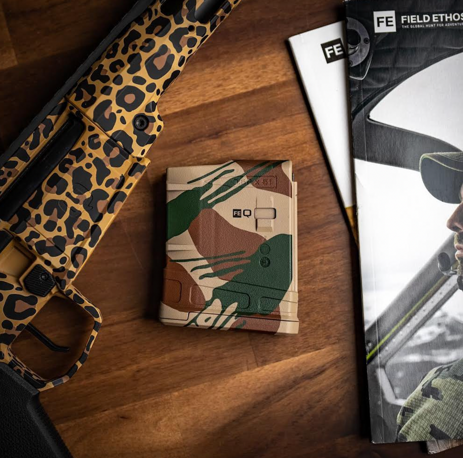 Rhodesian brushstroke camo Q x Field Ethos magazine sitting on a table next to a rifle and stack of magazines.